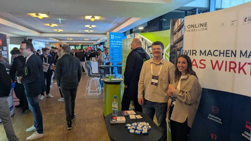 Zwei junge Mitarbeiter an einem Messestand der Online Rebellion mit einem Tisch voll Flyer und Werbegeschenke für interessierte.